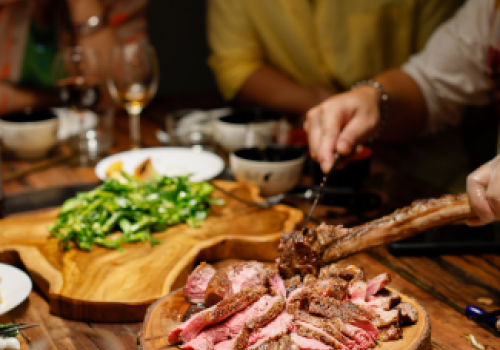 A group of people is dining, with sliced steak and green vegetables on wooden serving trays at the center of the table. Glasses and dishes are around.