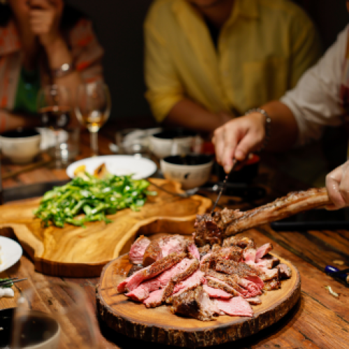 A group of people is dining, with sliced steak and green vegetables on wooden serving trays at the center of the table. Glasses and dishes are around.