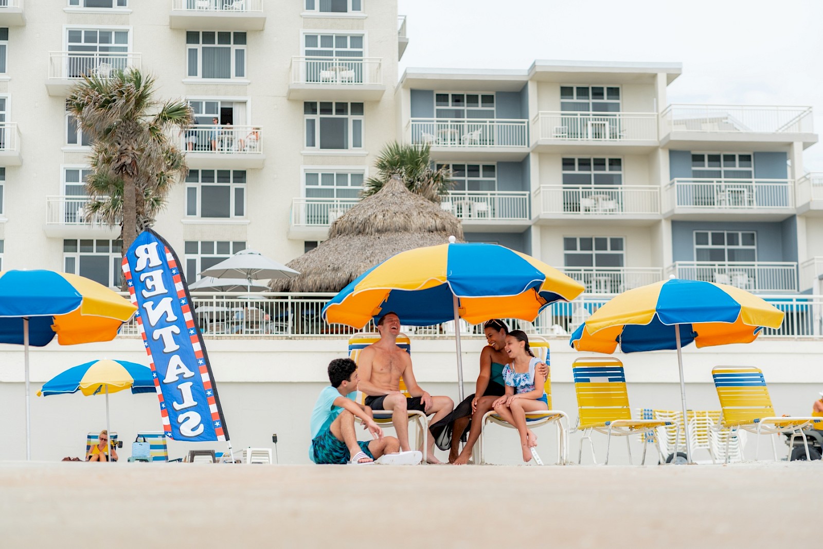 A group of four people enjoying a sunny day at the beach, sitting under colorful umbrellas with a building and palm trees in the background.