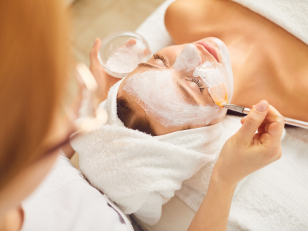 A person is receiving a facial treatment, with a mask being applied to their face using a brush, while wrapped in a towel.