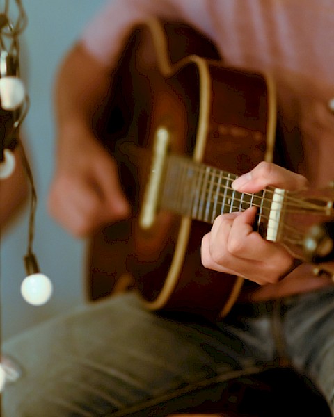 A person playing an acoustic guitar next to string lights in a cozy setting.