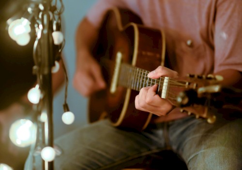 A person playing an acoustic guitar next to string lights in a cozy setting.