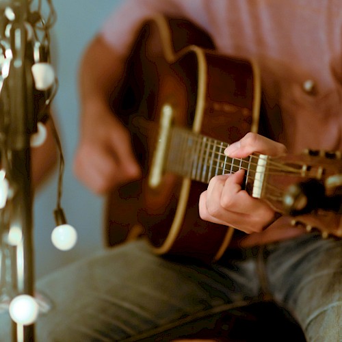 A person playing an acoustic guitar next to string lights in a cozy setting.
