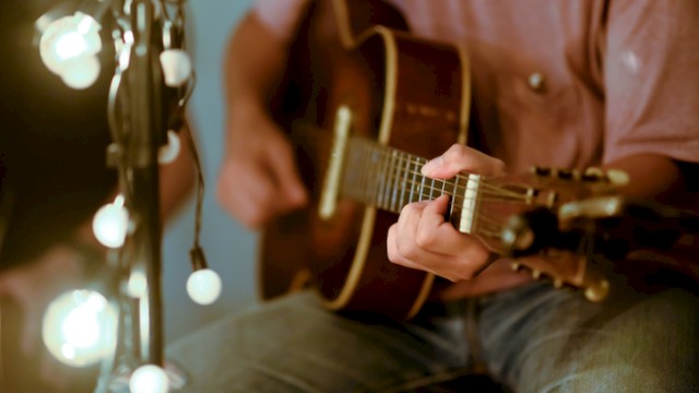 A person playing an acoustic guitar next to string lights in a cozy setting.