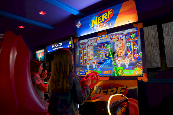 Two children play an arcade shooting game called Nerf Arcade, with colorful lights and game screens in a dark arcade room.