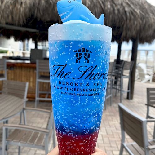 A hand holds a red and blue layered drink with a blue shark toy on top in a branded cup from 