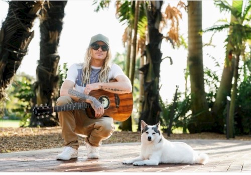 A man with long blonde hair, tattoos, sunglasses, and a cap sits with a guitar while a white dog with black markings relaxes on a sunny, tropical path.