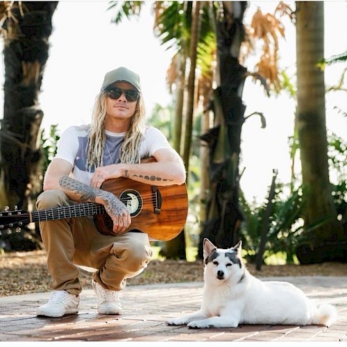 A man with long blonde hair, tattoos, sunglasses, and a cap sits with a guitar while a white dog with black markings relaxes on a sunny, tropical path.