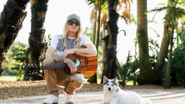 A man with long blonde hair, tattoos, sunglasses, and a cap sits with a guitar while a white dog with black markings relaxes on a sunny, tropical path.