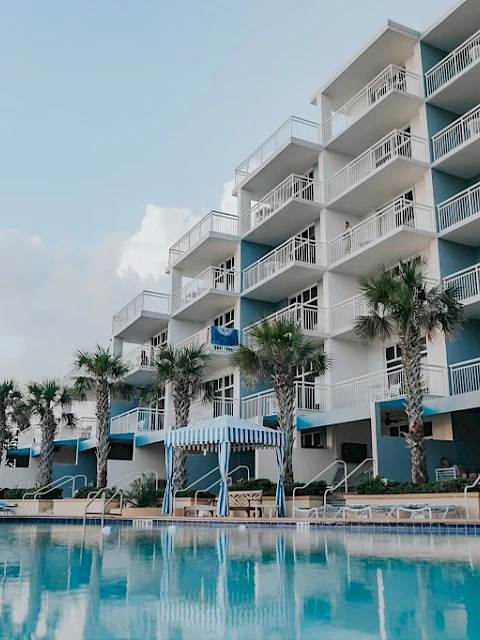A hotel with a pool and palm trees, balconies, and lounge chairs in a sunny, tropical setting.