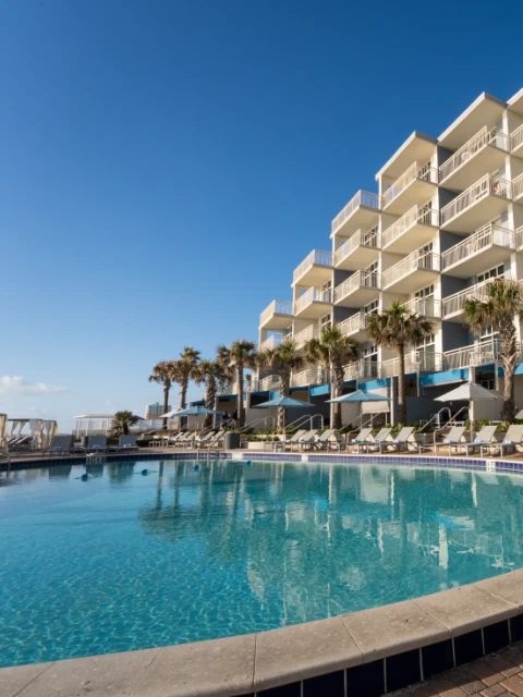 A hotel pool area with lounge chairs, umbrellas, palm trees, and a large white building in the background under a clear blue sky.