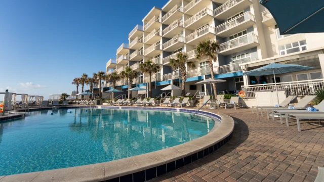A hotel pool area with lounge chairs, umbrellas, palm trees, and a large white building in the background under a clear blue sky.