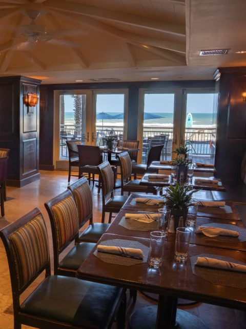 A cozy, well-lit restaurant with wooden furniture, large windows overlooking a beach, and outdoor seating beneath umbrellas.