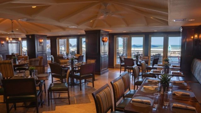 A cozy, well-lit restaurant with wooden furniture, large windows overlooking a beach, and outdoor seating beneath umbrellas.