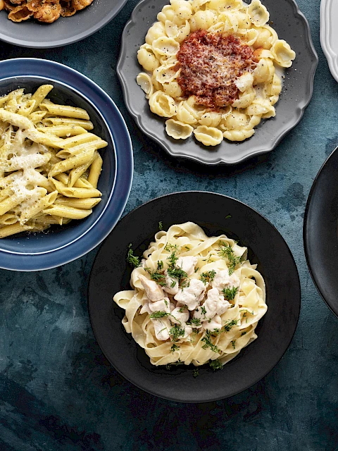 Various plates of different pasta dishes on a blue textured surface, including penne, tortellini, and spaghetti with toppings.