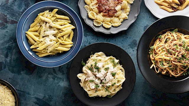 Various plates of different pasta dishes on a blue textured surface, including penne, tortellini, and spaghetti with toppings.