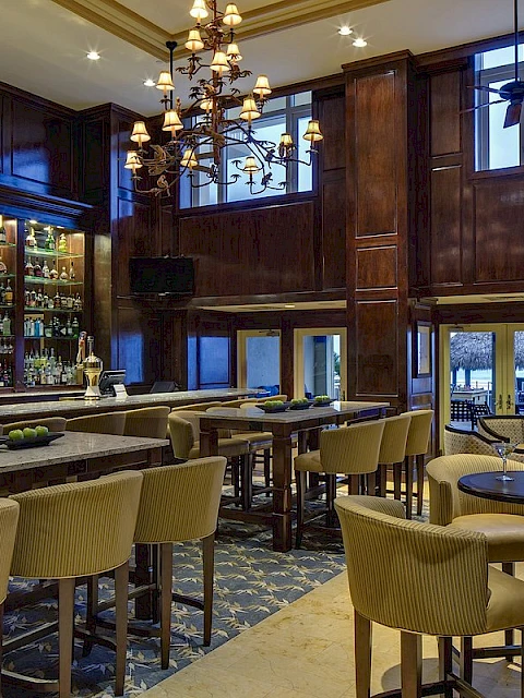 An upscale bar/restaurant interior with wooden paneling, high windows, and a view of a beach outside.