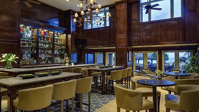 An upscale bar/restaurant interior with wooden paneling, high windows, and a view of a beach outside.