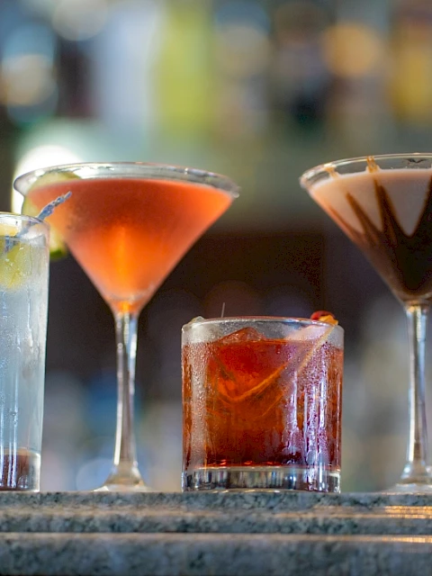 Four colorful cocktails are displayed on a bar counter, each with distinct appearances and garnishes, set against a blurred bar background.