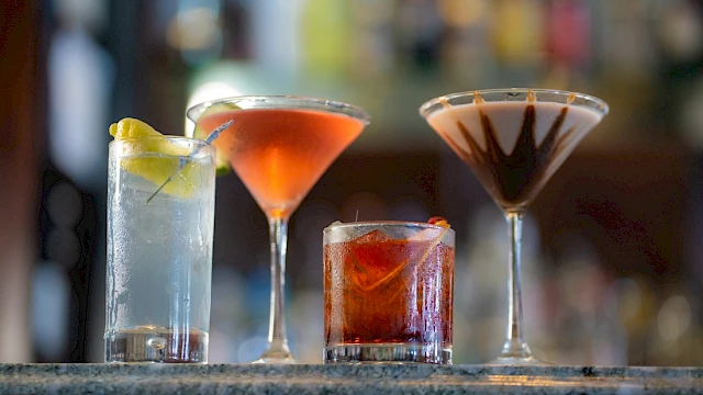 Four colorful cocktails are displayed on a bar counter, each with distinct appearances and garnishes, set against a blurred bar background.