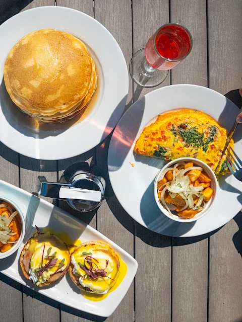 A top-down view of a meal including pancakes, an omelette, eggs Benedict, sweet potato fries, and drinks on a wooden table.