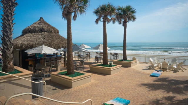 A sunny beachside scene with palm trees, a thatched hut, umbrellas, chairs, and a view of the ocean.