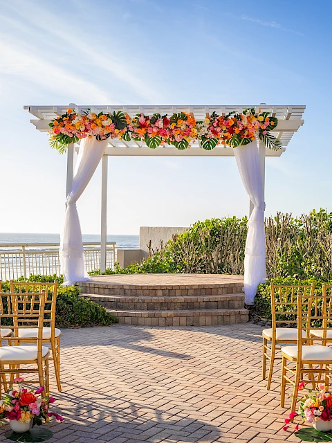A scenic seaside wedding altar with colorful flowers, golden chairs, and a clear sky over the ocean.