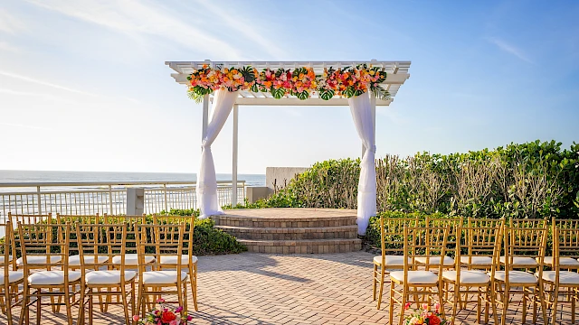 A scenic seaside wedding altar with colorful flowers, golden chairs, and a clear sky over the ocean.
