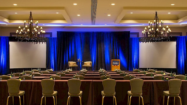 A conference room setup with chairs, tables, large screens, a podium, and chandeliers for an event or presentation.