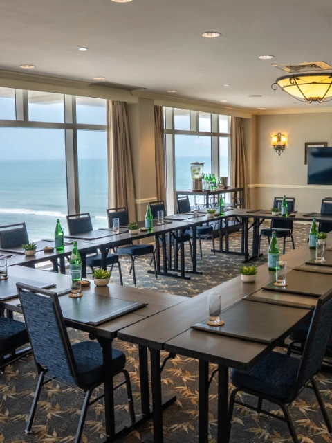 A conference room with a U-shape table setup, chairs, water bottles, glasses, and a view of the ocean through large windows.