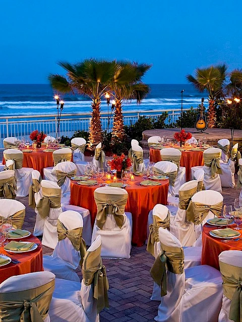 An outdoor beachside dinner setup with tables, chairs, candles, flowers, and a guitar on a stage against the ocean at dusk.