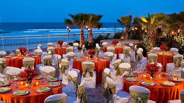 An outdoor beachside dinner setup with tables, chairs, candles, flowers, and a guitar on a stage against the ocean at dusk.