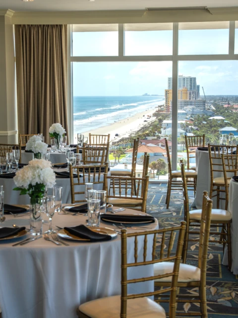 A beautifully set banquet room with round tables, white tablecloths, black napkins, and elegant gold chairs, overlooking a beach and ocean.