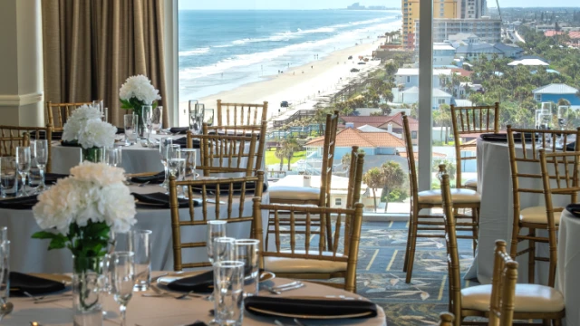 A beautifully set banquet room with round tables, white tablecloths, black napkins, and elegant gold chairs, overlooking a beach and ocean.