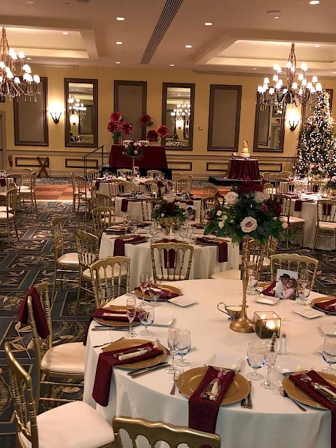 A beautifully decorated banquet hall with round tables, floral centerpieces, elegant table settings, and a Christmas tree.