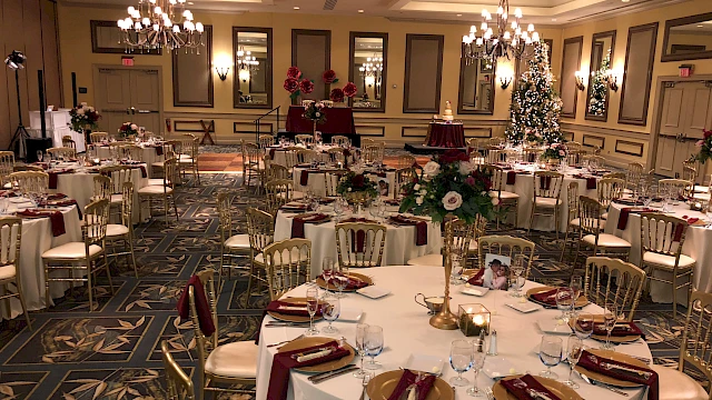 A beautifully decorated banquet hall with round tables, floral centerpieces, elegant table settings, and a Christmas tree.