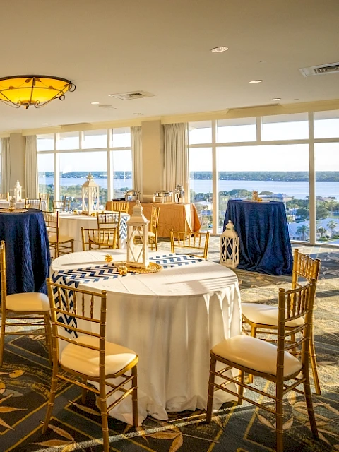 A spacious, well-lit banquet hall with elegant tables, chairs, and decor, overlooking a scenic waterfront view outside.