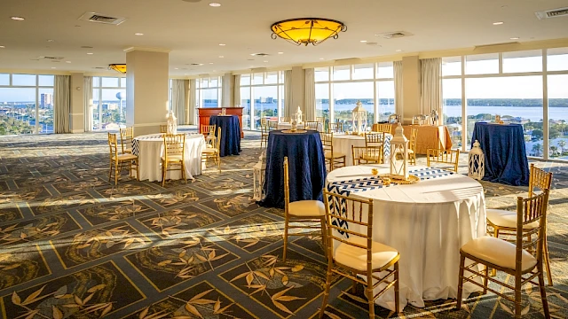 A spacious, well-lit banquet hall with elegant tables, chairs, and decor, overlooking a scenic waterfront view outside.