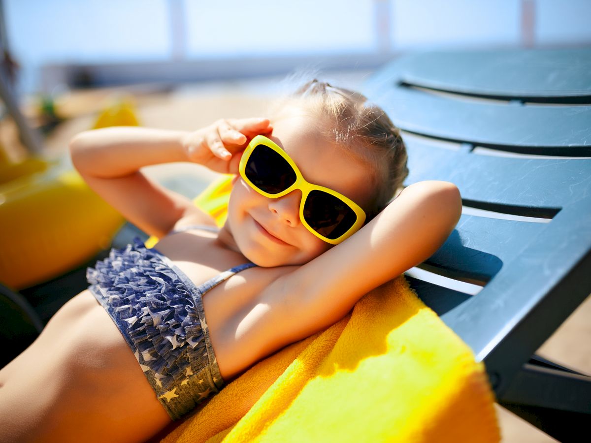 A young girl is lounging on a sunbed with a yellow towel, wearing yellow sunglasses and a swimsuit, smiling and enjoying a sunny day.