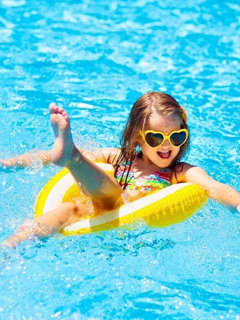 A joyful girl wearing sunglasses rides a yellow float in a swimming pool, enjoying sunny weather and summer fun.