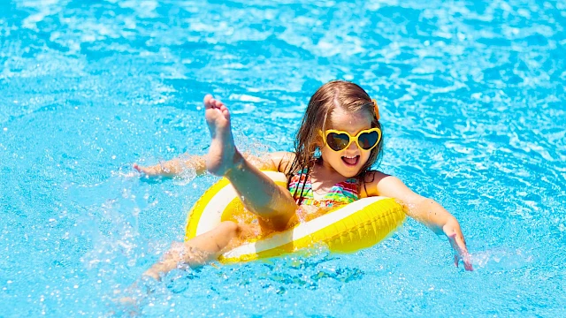 A joyful girl wearing sunglasses rides a yellow float in a swimming pool, enjoying sunny weather and summer fun.
