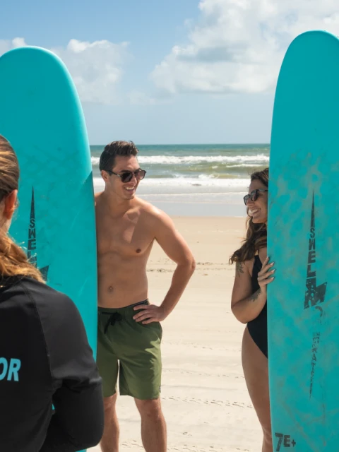 A surf instructor teaches two smiling students at a sunny beach, holding surfboards and enjoying their time by the ocean.