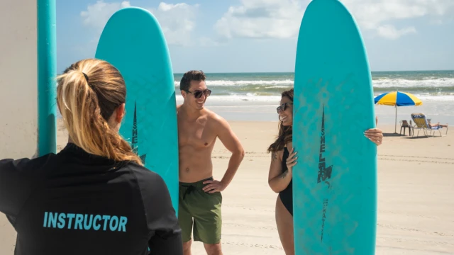 A surf instructor teaches two smiling students at a sunny beach, holding surfboards and enjoying their time by the ocean.