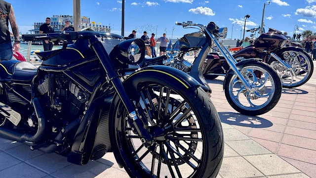 A lineup of custom and luxury motorcycles displayed outdoors on a sunny day at a bike show with people in the background.
