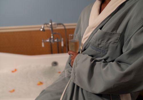 Person in a bathrobe holding a glass, standing beside a bubble-filled bathtub with rose petals. The faucet is visible in the background.