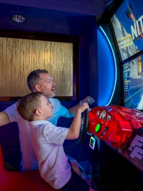 A father and son playing a high-speed racing arcade game together indoors.
