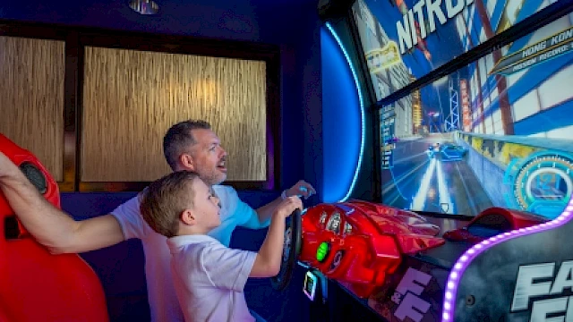A father and son playing a high-speed racing arcade game together indoors.