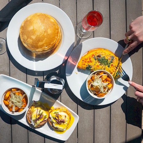 A table with breakfast food including pancakes, an omelette, bread slices, and drinks on a wooden table outdoors.
