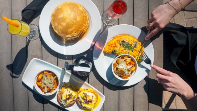 A table with breakfast food including pancakes, an omelette, bread slices, and drinks on a wooden table outdoors.