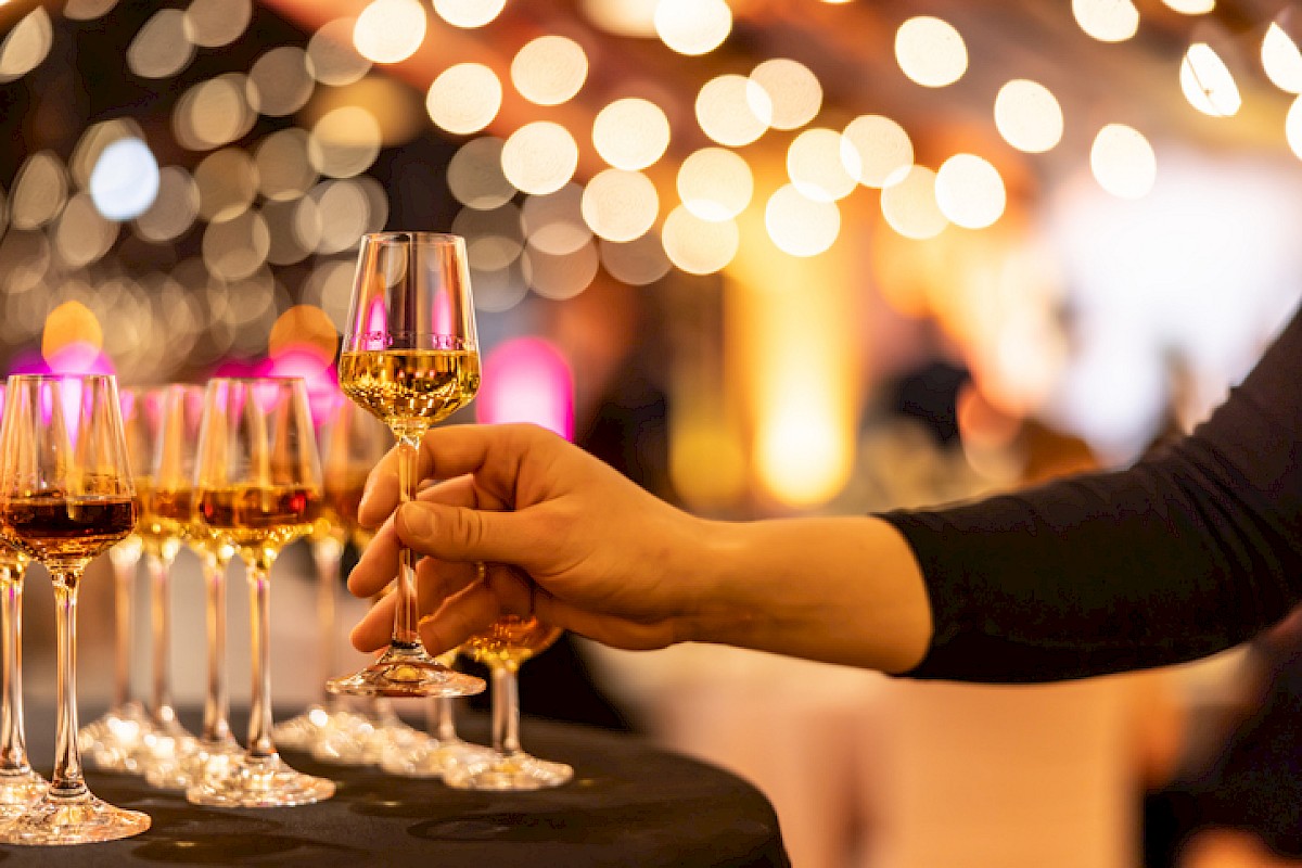 A person is holding a glass of white wine at an event or celebration with multiple glasses and warm, bokeh lights in the background.